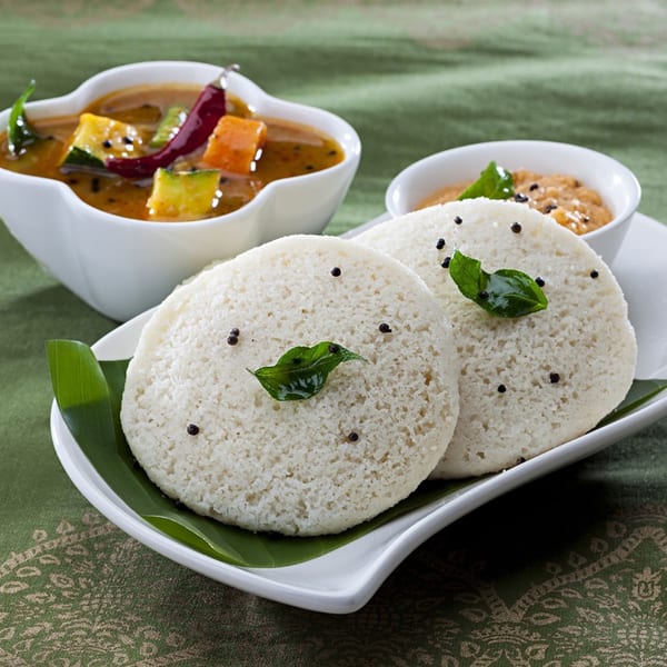 Two idlis garnished with curry leaves and peppercorns, served with sambar and chutney.
